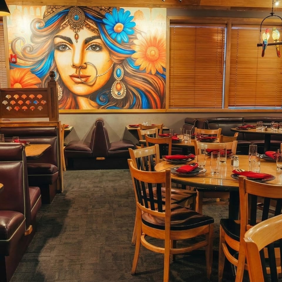 Warmly lit restaurant dining room with wooden tables, red napkins, leather booths, and a vibrant mural of an Indian woman.
