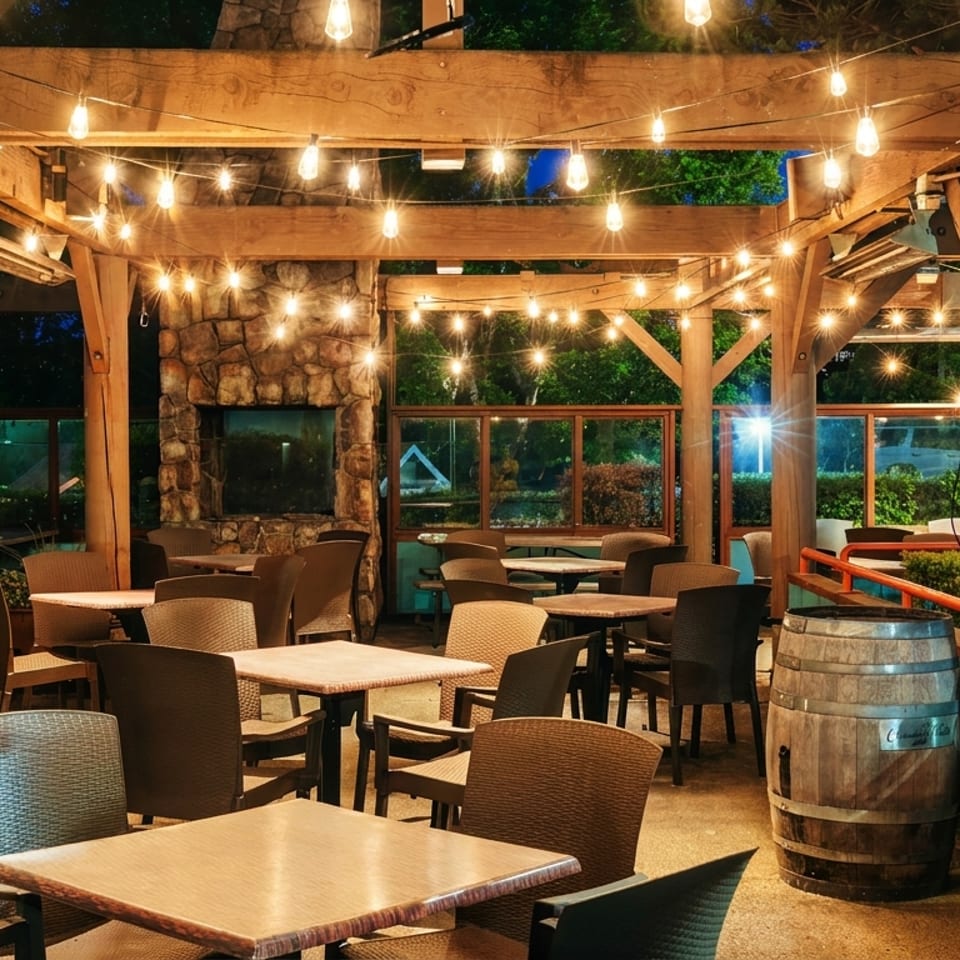 Outdoor restaurant patio with wooden beams, string lights, tables, chairs, barrel tables, and a stone fireplace at night.