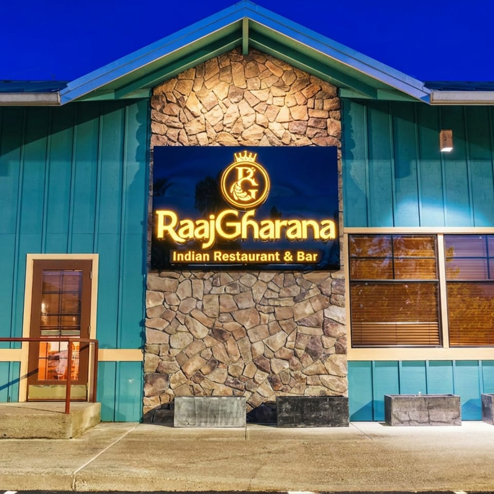 Nighttime exterior of RaajGharana Indian Restaurant & Bar, featuring teal siding, stone facade, illuminated sign, windows, and planters.